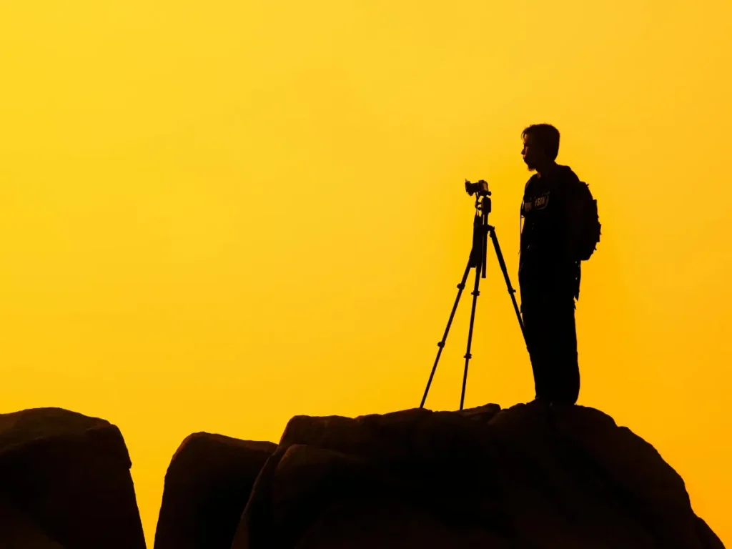 Silhueta de um fotógrafo no topo de uma montanha ao amanhecer com câmera e tripé - Curso de Fotografia Online: Quase Gratuito e de Alta Qualidade