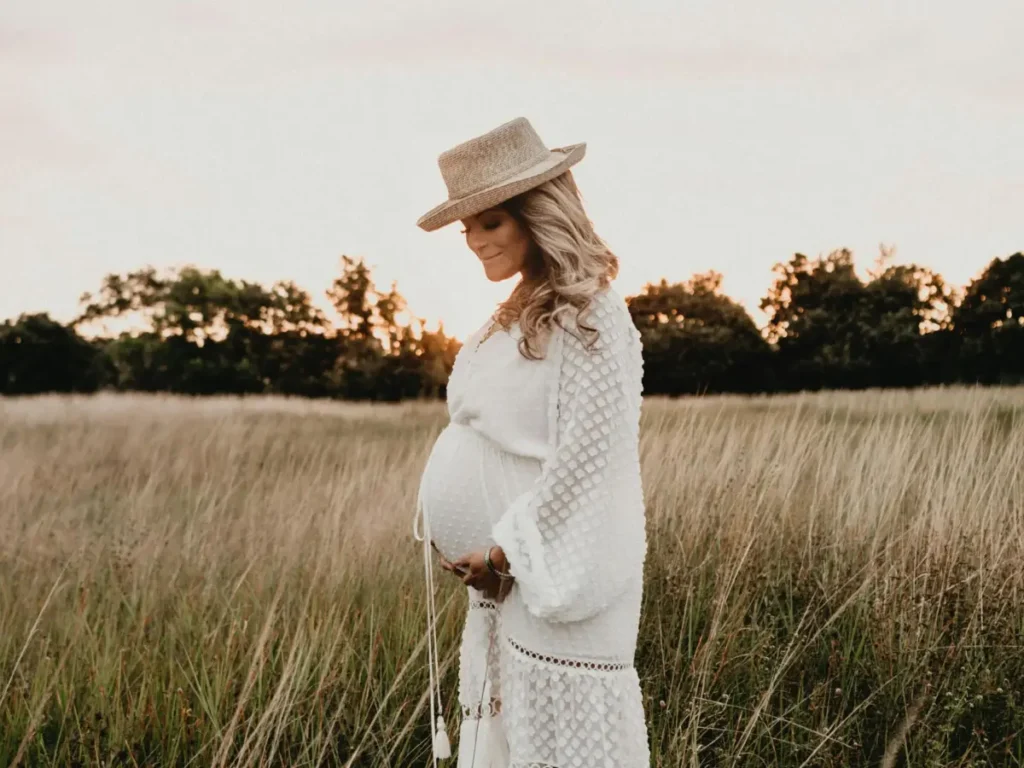 Gestante em um campo ao ar livre ao pôr do sol, vestindo um vestido branco e chapéu, representando o Curso de Fotografia para Gestantes